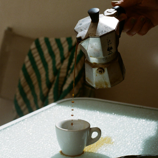 Person pouring coffee from a Moka pot into a white cup on a glass table.
